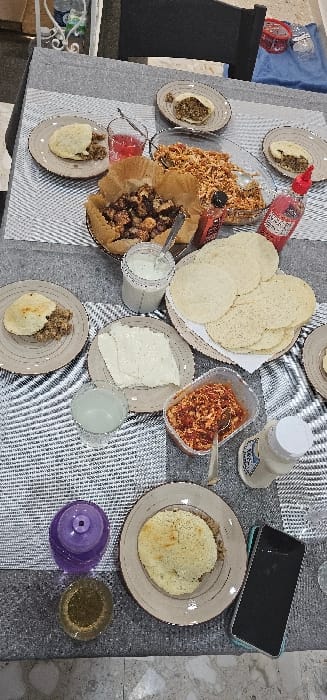 The image presents an overhead view of a dining table set with various prepared food items, situated within a residential environment in Birkirkara, Malta. The table surface is predominantly covered by a grey and white patterned placemat.
Multiple beige plates are arranged, each holding a round, flat, pale yellow bread, some of which are partially filled with a brown, textured substance. One plate contains several slices of white cheese. A stack of plain, round, pale yellow flatbreads is present on another plate, wrapped in white paper.
Central to the arrangement are a large clear glass bowl filled with shredded orange-red food and a brown paper-lined basket containing numerous dark, irregularly shaped food pieces. A clear plastic container holds a reddish, textured food item. A clear jar contains a white, creamy liquid, with a spoon inserted.
Beverages include a clear glass with red liquid, a clear glass with pale green liquid, and another clear glass with amber liquid. A purple glass or container is also on the table. Condiments include a red squeeze bottle and a white plastic bottle with a white cap. A smartphone with a black screen and a light blue/teal case rests face down near one of the plates.
In the background, a dark chair back is partially visible. Sections of a white patterned floor or wall are discernible on the left and bottom edges of the frame. A clear plastic bottle and a red plastic container are visible in the upper background. No individuals are present in the scene.