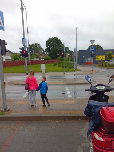 In Kuršėnai, Lithuania, on a cloudy day, a woman in a pink jacket and gray pants, and a boy in a blue jacket and black pants are holding hands and standing on a crosswalk. They are facing away from the camera, looking towards the street. A traffic light shows a red signal, indicating that pedestrians should not cross. The pavement is wet, suggesting recent rain. In the foreground, the rear of a red scooter covered with a blue tarp is visible. The background features a grey building with yellow signage, trees, and residential houses. A traffic light, pedestrian crossing signs, and streetlights are also present.