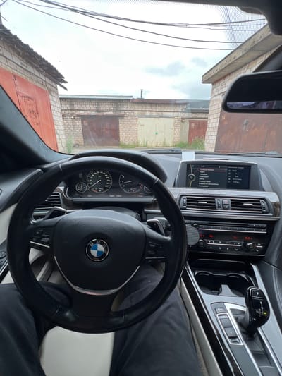 The image is a first-person view from the driver's seat of a BMW car parked in what appears to be a garage complex. The car's interior is visible, including the steering wheel with a BMW logo, dashboard with a digital display showing navigation and vehicle information, climate controls, and gear shifter. Outside the car, through the windshield, rows of garages with corrugated metal doors are lined up. The garages are constructed of brick and appear to be in a state of disrepair. The sky is overcast. The location is identified as Veteran-2, Russia.