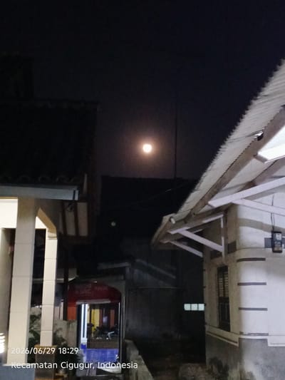 The image depicts a night scene with a full moon in a dark sky. In the foreground, there are parts of two buildings, one on the left with a white facade and pillars, and another on the right with a corrugated metal roof supported by wooden beams. The left building has a visible entrance with signage that is partially obscured but appears to be in red and white. The right building has a window with dark shutters and some electrical conduit attached to the exterior. The overall environment suggests a residential or semi-commercial area. The date stamp in the lower left corner reads "2026/06/29 18:29" and the location is identified as "Kecamatan Cigugur, Indonesia." There are no people visible in the image.