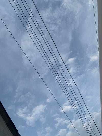 An upward-angle outdoor shot captures a sky filled with scattered, white, cumulus clouds against a pale blue backdrop. Several dark, thin electrical wires are diagonally strung across the frame, from the top left to the bottom right. In the lower left corner, the dark, corrugated metal roof of a building is visible, angled upwards. The right edge of the frame shows a sliver of a white, vertical structure, possibly a wall or pillar. The scene is set in Elelenwo, Nigeria.