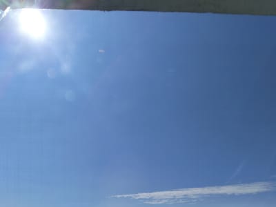 The image is a photograph of a clear blue sky with the sun prominently visible on the left side, creating a lens flare effect. There are a few faint circular blurs, possibly dust or water droplets on a lens or window, scattered across the sky. A thin strip of white clouds is visible near the bottom of the frame. The top edge of the image shows a dark, textured surface, which could be a building overhang or a similar structure. The scene appears to be an outdoor environment, likely taken during daylight hours. No people or specific city landmarks of Bulaev, Kazakhstan are identifiable in the image.