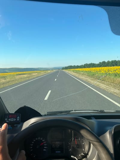 The image is taken from the interior of a vehicle, looking out through the windshield at a rural road. The road stretches into the distance, lined on the right by a large field of sunflowers and on the left by rolling hills. The sky is clear and blue.

Inside the vehicle, a steering wheel is visible in the foreground. A dashboard displays various instruments, including a speedometer. A device with buttons and a digital display, possibly a cruise control unit, is mounted to the left of the steering wheel. A person's hand is visible on the left side of the frame, interacting with the device.

The scene appears to be in Cimișlia, Moldova, indicated by the presence of sunflower fields, which are common in the Moldovan countryside, and the context provided. The time appears to be morning or early afternoon, given the bright sunlight and clear sky.