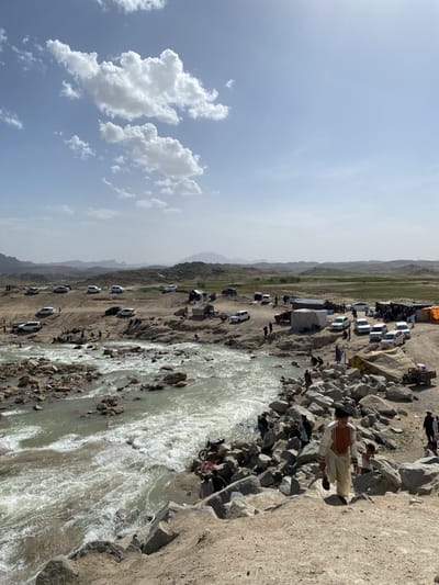 The image depicts a vibrant scene in Kim, Afghanistan, showcasing a river flowing through a rocky landscape. The river's waters are turbid, suggesting a strong current. On both banks of the river, people are gathered, some in the water, others on the rocks or the shore. Several vehicles are parked nearby, indicating a popular gathering spot. Tents and shelters are erected on the land, possibly for picnics or gatherings. One individual is seen walking down a rocky embankment towards the river, wearing traditional clothing. The sky is partly cloudy, with visible mountain ranges in the distance. The overall environment suggests a natural outdoor recreational area.