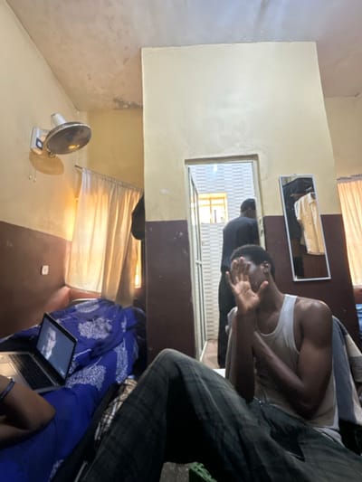 The image depicts an interior room located in Ado-Ekiti, Nigeria. The room features a two-toned wall design with a cream-colored upper section and a dark brown lower section. The ceiling shows evidence of wear or staining.

On the left wall, a white wall-mounted oscillating fan is positioned above a window draped with light-colored curtains, through which bright natural light illuminates the area. On a surface below the window, covered with blue patterned fabric, a silver laptop computer is open, displaying a screen that shows the face of a man wearing glasses.

In the mid-ground, a dark-skinned male subject is seated, wearing a light-colored tank top and dark plaid trousers. His right hand is raised, obscuring his face.

An open doorway in the center-right leads to an adjacent space with light-colored, horizontally striped walls. A second dark-skinned male subject is visible from the back within this doorway, wearing a dark long-sleeved garment and dark trousers. To the immediate right of the doorway, a rectangular mirror is mounted on the wall, reflecting or displaying a light-colored shirt. A green object is partially visible on the floor in the lower right foreground.