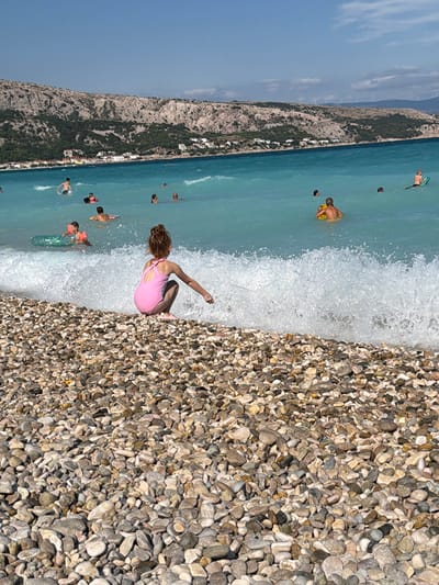 In the city of Općina Baška, Croatia, on a sunny day, a young girl with brown hair pulled up in a bun is crouched on a pebble beach, facing the turquoise sea. She is wearing a pink swimsuit. In the background, several people are swimming and playing in the water. A large wave is crashing on the shore near the girl. The shoreline is covered in various sizes of grey and brown pebbles. Beyond the beach, a rocky, tree-covered hill rises, with a town nestled along its base. The sky is clear and blue, with a few wispy clouds.