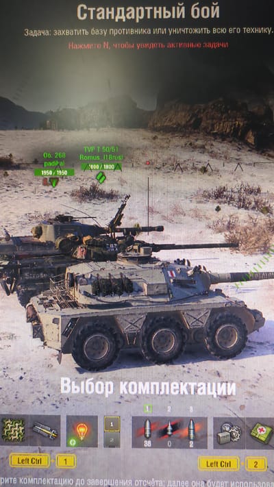 The image displays a scene from a video game, likely a tank combat simulation. The user interface elements are prominent, showing game objectives and controls. The top of the screen displays "Стандартный бой" (Standard Battle) and a description of the objective: "Задача: захватить базу противника или уничтожить всю его технику." (Objective: Capture the enemy base or destroy all their equipment.) Below this, it instructs the player to "Нажмите N, чтобы увидеть активные задачи" (Press N to see active tasks).

In the game's environment, two armored vehicles are visible. The front vehicle is a wheeled tank with multiple wheels and a turret. The second vehicle, positioned behind and partially obscuring the front one, appears to be a tracked tank. The environment is a desolate, sandy landscape with sparse vegetation and rocky formations in the background, suggesting a desert or arid setting.

The bottom part of the image shows a "Выбор комплектации" (Loadout Selection) menu. It displays icons for various items, including camouflage, equipment, a consumable item (represented by a lightbulb), and ammunition. Specific ammunition types are shown with quantities: 38 of one type, 0 of another, and 2 of a third. Control prompts like "Left Ctrl + 1" and "Left Ctrl + 2" are visible, indicating player input options.

There is no indication that this image is related to the city of Yeni Surakahny, Azerbaijan. The content is purely from a digital simulation.