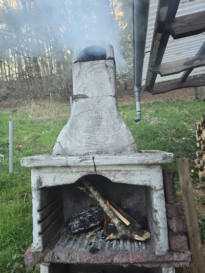 An outdoor, light grey concrete barbecue or fireplace with a chimney is captured in a daytime scene in Rotenburg an der Fulda, Germany. A fire is actively burning inside the firebox, with several logs and embers visible, indicating a recent ignition or ongoing use. A significant plume of white smoke billows out from the chimney opening, rising into the air.
The concrete structure shows signs of age and use, with some visible cracks and discoloration. It is situated on a patch of green grass. In the background, a stack of cut firewood is neatly piled on the ground, suggesting the fuel source for the fire. Further into the background, trees with mostly bare branches are visible. In the upper left corner of the image, part of a wooden structure, possibly a shed or pergola, with a translucent corrugated roof, is present. A grey pipe, possibly a drain or vent, runs horizontally across this section. No people are visible, and there is no discernible text in the image.