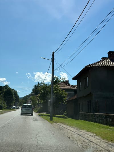 In Beli Osam, Bulgaria, a light blue car travels down a paved road on a sunny day. The car is in the right lane, moving away from the viewer, with trees lining the left side of the road and houses and more trees on the right. The sky is a clear blue with a few white clouds. Power lines are visible overhead, supported by a utility pole on the right side of the road. The road has some cracks and patches. A low stone wall runs along the side of the road in front of a house.