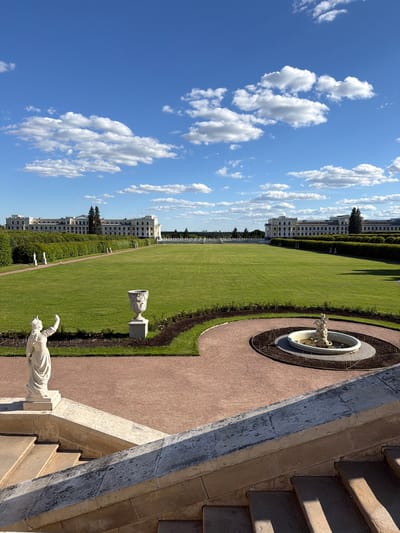 The image depicts a grand estate in Arkhangel'skoe, Russia, under a bright blue sky with scattered white clouds. In the foreground, a large stone staircase leads down to a manicured garden. To the left, a classical white statue of a female figure stands on a pedestal, its right arm raised. Directly ahead, a vast expanse of perfectly mown green lawn stretches towards a long, white neoclassical building. To the right of the lawn, a circular fountain with a white statue at its center is surrounded by dark soil and low-lying plants. The building, which appears to be a palace or manor, is symmetrical with rows of windows and a central colonnade. A neatly trimmed hedge runs along the perimeter of the lawn, separating it from pathways and more statues. The overall impression is one of formal elegance and vastness.