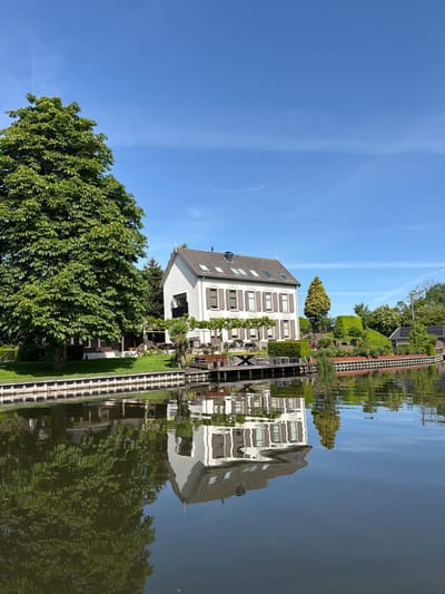 The image shows a house with a white facade and brown shutters, reflected in a canal. A large tree with green foliage stands near the house. A narrow walkway with a black and white checkered pattern extends along the canal, also reflected in the water. The sky is blue with faint clouds. The photo is taken in Leerdam, Netherlands, given the architectural style and canal.