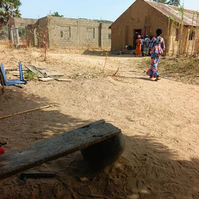 This media file depicts an outdoor scene in what appears to be a rural or semi-urban setting, likely in West Africa, with "Belnin, Nigeria" provided as a potential location context. The ground is predominantly sandy, with sparse, dry vegetation and scattered items.
In the foreground, a rudimentary structure is visible, consisting of a weathered wooden plank resting on a large, dark, rounded object, possibly a pot or stone, serving as a makeshift bench or table. Nearby, two blue plastic chairs are overturned or discarded, and a bamboo-like stick lies on the sand.
The midground features two distinct buildings. To the left, there is a partially constructed or unfinished building made of grey cinder blocks, featuring several window openings without frames. To the right, an older, more traditional house stands, constructed from reddish-brown mud or earth walls and topped with a corrugated iron roof.
An activity appears to be centered around the entrance of the mud house. A group of women, dressed in vibrant, patterned traditional West African attire, are gathered at the doorway, some seemingly entering or exiting. A prominent figure, a woman in a bright pink, blue, and black patterned dress, walks away from this group towards the right side of the frame, her back to the viewer, suggesting movement across the sandy area.
The time of day is bright and sunny, indicated by the clear sky and sharp shadows cast on the ground, suggesting midday or early afternoon in a dry climate. There are no discernible emotions conveyed by the distant figures. No visible text is present in the image. The scene captures a blend of traditional architecture and ongoing development.