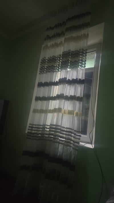 A sheer curtain with horizontal bands of dark gray, light gray, and yellow zig-zag patterns hangs in front of a window. The window frame is white, and the glass reveals the muted green color of another surface outside. The curtain is illuminated from behind, casting subtle shadows. To the left of the window, a portion of a dark, rectangular object is visible against a green wall. The room appears dimly lit, with a dark floor in the foreground. A thin black wire runs from the window area down the green wall. The overall scene suggests an interior space in Yakkabog, Uzbekistan.