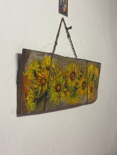 A framed painting of sunflowers on a wooden plank hangs on a white, textured wall. The painting is in the style of Van Gogh, with thick brushstrokes and vibrant yellow petals. The plank is oriented horizontally and is suspended by a twisted rope hanger. Above the painting, a small religious icon depicting the Virgin Mary and child is affixed to the wall. The lighting is natural, suggesting an indoor setting. The environment is a domestic interior in Shipkovo, Bulgaria, indicated by the presence of the religious icon, commonly found in Orthodox homes.
