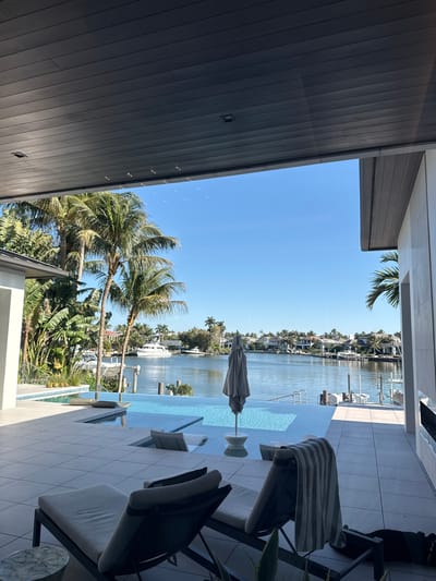 This image depicts a luxurious outdoor waterfront property in Naples, United States of America, under a clear blue sky.
The scene is viewed from a covered patio with a dark grey slatted ceiling. The patio floor consists of large, light-colored square tiles. In the immediate foreground, two dark-framed lounge chairs with light cushions are positioned facing the water. A striped towel (grey and white) is draped over the rightmost lounge chair. A small, round, light-colored side table with a patterned surface is next to the left chair, and a dark planter with a green plant is in the bottom right.
An infinity-edge swimming pool with light blue water is central to the image, situated directly off the patio. Submerged ledges or seating areas are visible within the pool. A folded grey beach umbrella stands upright within a shallow section of the pool.
Beyond the pool is a calm, wide waterway or canal. On the opposite side of the waterway, several boats, including a prominent white motor yacht, are docked. The shoreline is lined with lush tropical vegetation, primarily various palm trees, interspersed with large, upscale residential homes with modern architecture. The sky is bright and clear, indicating full daylight. No people or animals are visible, and no actions or interactions are depicted.