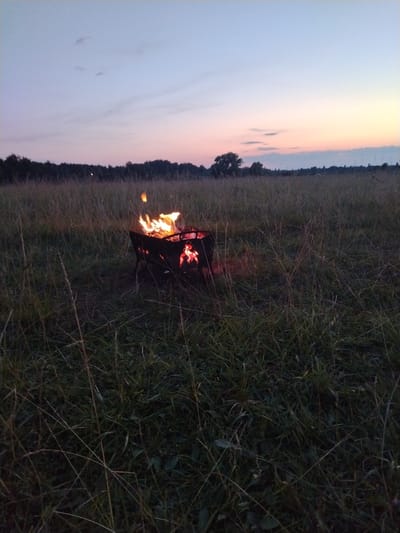 In the city of Kriukovskoe, Russia, an outdoor scene unfolds at dusk. A portable metal fire pit, filled with burning wood and embers, is situated in a field of tall grass. The flames are vibrant orange and yellow, casting a warm glow on the surrounding vegetation. The sky above displays a gradient of colors, transitioning from a pale blue at the top to soft pinks and yellows near the horizon, indicative of a sunset or twilight. A line of trees can be seen in the distance, bordering the field. The overall atmosphere is tranquil and natural, with no people or animals visible in the frame.
