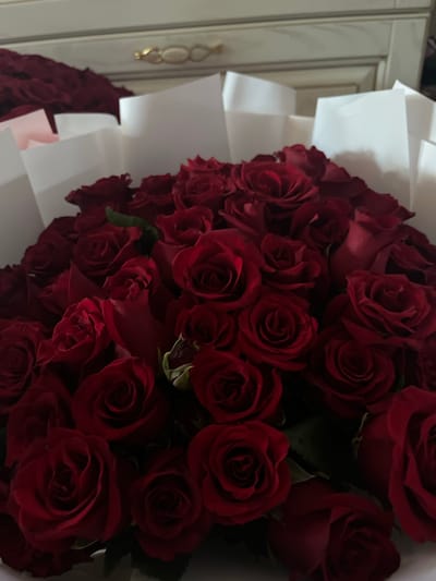 The image shows a close-up of a large bouquet of deep red roses, expertly arranged and partially wrapped in white paper. The roses are the primary subject, with their velvety petals and classic bloom shape clearly visible. Some roses are in full bloom, while others are still developing, showcasing a variety of stages. A few green leaves and stems are interspersed among the flowers, adding depth and texture to the bouquet.

The bouquet is presented against a backdrop that appears to be the interior of a room. To the left of the flowers, part of a piece of furniture, possibly a dresser or cabinet, can be seen. It is light-colored, with decorative molding. A decorative gold-toned handle or knob is also visible on this furniture. The lighting suggests an indoor setting, likely during the day, with soft shadows indicating ambient light rather than direct sunlight. The overall mood conveyed by the deep red roses is one of romance, elegance, and passion. There is no visible text in the image. The context provided places this scene in Pănășești, Moldova.