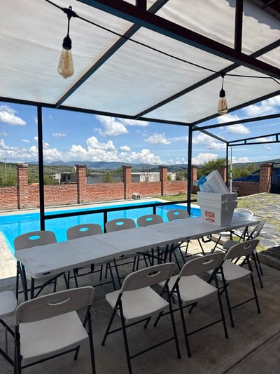 This image depicts an outdoor covered patio area with a swimming pool in the background. A long, white folding table is set up with multiple white plastic chairs around it. On the table, there is a white cooler with the OXXO logo. Beyond the patio, a bright blue swimming pool is visible, bordered by a brick wall. In the distance, hills and mountains are seen under a blue sky with scattered clouds. The overall scene suggests a relaxed outdoor gathering or event space. The location is in San Baltazar Torija, Mexico.