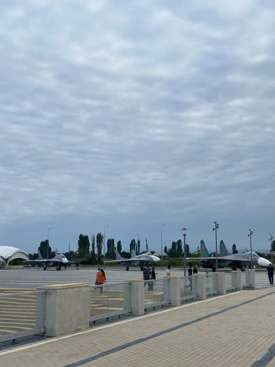 The image captures an outdoor scene with several military jets parked on a tarmac. The sky is overcast with gray clouds. In the foreground, there is a paved walkway and a low concrete barrier with a glass railing. Behind the barrier, there are stairs leading down. Several people are visible, some standing near the barrier and others walking on the tarmac. In the middle ground, two fighter jets are prominently displayed. One jet is gray with a white nose cone, and the other is a darker gray with teal accents on its tail fins. Further back, more trees and structures are visible, suggesting an airfield or an aviation museum. The location is identified as Arablyar, Russia.