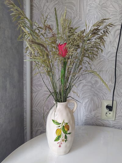 A still life photograph taken indoors displays a white ceramic vase with a lemon and blossom design. The vase, positioned on a white table, holds a bouquet of dried grass and a single bright pink tulip. Behind the vase, a light-colored wall with a leaf-patterned wallpaper is visible. To the right of the vase, a white electrical outlet with a black cord plugged in is mounted on the wall. A white vertical pipe is also present on the left side of the image. The overall scene suggests a domestic setting. The location is Gusev, Russia.