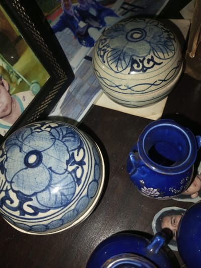 A collection of ceramic objects sits on a dark surface. Two round, light-blue and white boxes with floral patterns are visible. A small, dark-blue pot with tiny handles and floral decorations is also present. Two more dark-blue objects, likely cups, are partially visible at the bottom of the image. Fragments of photographs with faces are scattered around the items. A framed photograph is positioned to the left, partially obscuring a photo behind the boxes.