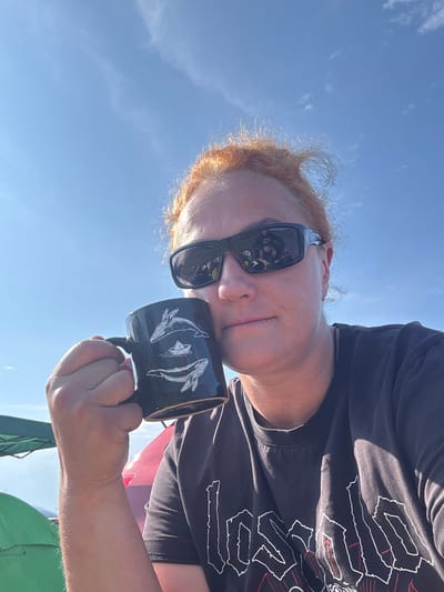 A woman with auburn hair and sunglasses is taking a selfie outdoors under a clear blue sky. She is holding a black mug decorated with white drawings of whales and a paper boat. She is wearing a dark t-shirt with a white graphic design. In the background, parts of green and red objects, possibly tents and a vehicle, are visible. The scene appears to be in an outdoor recreational area.