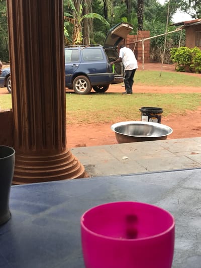 The image shows an outdoor scene in Otukpa, Nigeria. A dark blue hatchback car is parked on a grassy and dirt area, with its rear door open, and a person is leaning into the car's trunk. A shiny, silver-colored basin or bowl sits on a dirt patch next to a darker, bucket-like container. In the foreground, a brown, ribbed, cylindrical structure, possibly a column or post, is partially visible, along with a pink plastic cup or container on a gray or blue surface. The surrounding environment includes trees, foliage, and what appears to be a brick structure in the background.