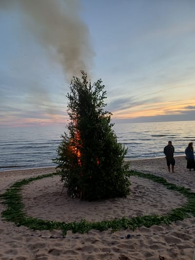 The image depicts a beach scene at sunset in Vārzas, Latvia. In the foreground, a dense green bush is ablaze, with flames visible through its branches and smoke billowing upwards. Two people are standing near the water's edge, one further back and appearing to observe the scene. The beach is sandy, with a curved border of green vegetation. The ocean is calm with gentle waves, and the sky is painted with the soft colors of sunset, transitioning from a pale blue to hues of orange and pink. The overall mood is serene, with a touch of dramatic intensity from the burning bush. The time of day is clearly sunset, indicated by the sky's colors. No text is visible in the image.