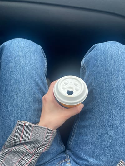 The image shows a person sitting in what appears to be a car, as indicated by the dark, smooth surface visible on the left. The person is wearing blue jeans and a blazer with a houndstooth pattern in black, white, and red. They are holding a disposable coffee cup with a white plastic lid, the lid bearing several small indentations and a larger opening for drinking. No specific location in Maribor, Slovenia, can be determined from this image. There are no other people or interactions visible.