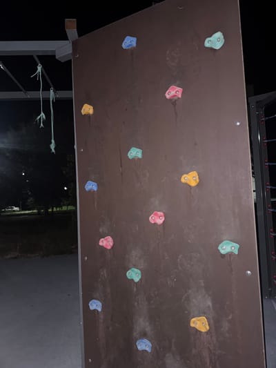 A climbing wall is pictured in Egorlykskaya, Russia. The wall is made of dark brown material and is covered with various colored climbing holds. There are blue, yellow, red, and green climbing holds attached to the wall. The scene is outdoors and appears to be at night, as indicated by the dark environment and visible streetlights in the background. Part of a metal structure with ropes hanging from it is visible on the left side of the image, suggesting this may be a playground or outdoor recreation area. The ground below the climbing wall is paved.