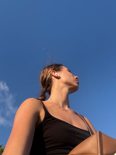 A low-angle shot captures a young woman from the shoulders up, set against a clear, bright blue sky with faint wisps of clouds. She is facing to the right and looking upwards, her eyes closed and her lips slightly parted. The woman has light brown hair pulled back in a ponytail and is wearing white wireless earbuds in her right ear. A delicate necklace with small pearls adorns her neck. She is wearing a black tank top, and a portion of a tan leather bag is visible on her right side. A small mole is present on her right shoulder. The lighting suggests it is daytime, possibly late afternoon due to the warm glow on her skin. The image appears to be taken outdoors, given the vast expanse of sky and the faint hint of greenery in the far lower-left corner, suggesting a natural or park-like setting within the city of Selongbelanak, Indonesia.