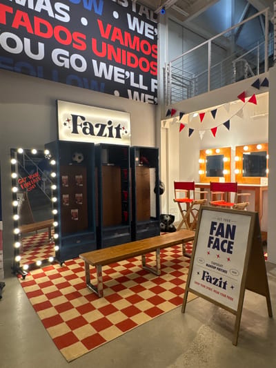 In a well-lit indoor space, likely a retail or promotional environment, the scene is dominated by a locker room-style setup. A row of dark blue lockers stands against a gray wall, with a wooden bench in front of them. To the left, a large mirror with vanity lights reflects the space, displaying a sign that reads "GET YOUR FAN FACE ON!". Above the lockers, a backlit sign with the word "Fazit" is illuminated. A red and beige checkered rug covers the floor in front of the lockers and mirror.

Further into the space, a row of makeup stations with lighted mirrors and red director's chairs suggests a beauty or performance-related theme. A wooden table supports these stations. In the background, a mezzanine level with metal railings is visible. Decorative triangular flags in red, white, and blue are strung across the upper portion of the room.

A freestanding A-frame sign stands prominently in the foreground, angled towards the viewer. The sign features bold blue text that says "GET YOUR FAN FACE" above smaller text indicating "TEMPORARY MAKEUP PATCHES" and "HAIR," along with the brand name "Fazit" and a tagline.

The overall impression is a curated, themed display designed to engage visitors. The lighting is bright, creating a clean and somewhat theatrical atmosphere. The presence of sports-related items like a soccer ball and pennants, combined with makeup stations and motivational signage, suggests a connection to sports fandom and fan engagement. The image appears to have been taken at eye level, capturing the display from a slightly elevated perspective. The location in Marina del Rey, United States, is not explicitly visible but is provided as context.
