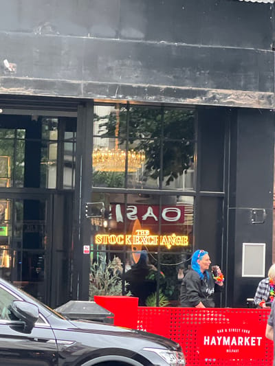 In Belfast, Northern Ireland, a woman with bright blue hair, wearing a black hoodie and a rainbow-colored lei, drinks from a dark beverage. She is positioned behind a bright red barrier at a venue labeled "HAYMARKET BELFAST," which also states "BAR & STREET FOOD." Behind her, a neon sign glows "THE STOCK EXCHANGE." The setting appears to be a city street, with a dark-colored car in the foreground. The building has large windows reflecting a chandelier and interior plants, suggesting an indoor space with a sophisticated ambiance contrasting with the vibrant street presence.