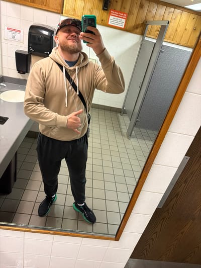 A selfie taken in a bathroom features a man wearing a beige hoodie, black sweatpants, and sneakers with green accents. He is holding a smartphone with a green case and is positioned in front of a mirror. The bathroom has white tiled walls and floors, and a brown wooden ceiling. Visible in the background are bathroom fixtures such as a sink, soap dispenser, and a paper towel dispenser. A sign on the wall reads "WASH HANDS". The location is likely Downers Grove, United States of America.