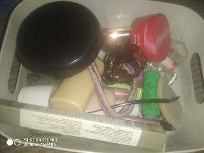 The image captures an overhead view of a white plastic storage container, located in Lumajang, Indonesia, filled with an assortment of personal care and cosmetic items. The container is rectangular with rounded corners and features vertical slot-like cutouts along its sides.

Essential details of the contents include:
*   A large, black, circular lid or compact case is positioned in the upper-left quadrant.
*   In the upper-right, a red, round plastic jar bearing the label "Kelly Pearl Beauty" is visible.
*   A dark brown translucent claw hair clip is situated near the red jar.
*   A light yellow opaque plastic bottle and a smaller white opaque plastic bottle are centrally located.
*   A pink and white striped elastic hair tie is intertwined among various items.
*   A small, bright green plastic tube is partially visible in the middle-right.
*   A round, light brown makeup sponge or puff rests on the right side of the container.
*   A thin, black, curved applicator tool lies across the green tube and the makeup sponge.
*   A rectangular, light-colored cardboard box, featuring small printed text, is placed horizontally across the bottom of the container.
*   Additional smaller, indistinct plastic bottles and tubes of various colors are interspersed throughout the container.

No individuals are present in the scene, and no specific actions or interactions are depicted. The image displays a watermark in the bottom-left corner stating, "SHOT ON REDMI 7 AI DUAL CAMERA."