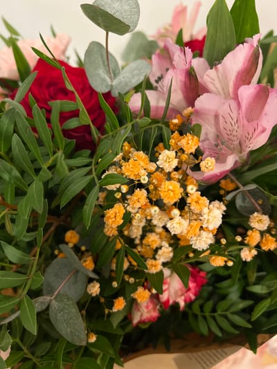 The image presents a close-up, high-angle view of a dense, mixed floral bouquet. The primary subjects are various flowers and foliage, arranged tightly together.
Prominently featured are:
* A vibrant red rose, partially open, located on the left side, slightly obscured by green leaves.
* Several pink Alstroemeria (Peruvian Lilies) with visible dark speckles on their petals, concentrated on the upper right and center-right.
* Clusters of small, globular orange-yellow flowers, resembling dried or dyed Gypsophila (baby's breath), distributed throughout the center and lower right, with some lighter cream-colored flowers intermingled.
* Various green foliage, including larger, rounded, pale blue-green leaves consistent with eucalyptus, positioned towards the top center and lower left.
* Numerous slender, dark green, elongated leaves with a distinct vein pattern, forming a significant part of the bouquet's structure, visible on the left and central areas.
* Hints of other pink or reddish flowers, possibly carnations or similar, are visible at the bottom and lower right, partially concealed by the other elements.
The background is uniformly light and out of focus, suggesting an indoor setting. No people or specific actions are depicted. The bottom edge shows a slight blur, indicating the bouquet might be resting on a light-colored surface or wrapped in light-toned material.
The image content does not provide any visual information to determine the specific location in Vilanova I La Geltrú, Spain.