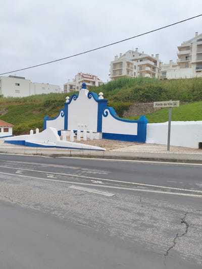 The image captures a street scene in Nazaré, Portugal. In the foreground, a gray asphalt road with visible cracks and white lane markings stretches across the frame. To the right, a sidewalk with light-colored paving stones leads to a street sign with text in Portuguese, "Fonte da Vila", and a symbol that appears to represent a fountain.
Behind the sidewalk is a decorative white and blue structure that appears to be a public fountain or monument, with curved blue accents and a central white section. This structure is situated on a grassy incline. Above the fountain, several multi-story white residential buildings with beige trim are visible, overlooking the scene. The sky is overcast and a dark cable is stretched across the upper portion of the image. A small, red-roofed building with brown window frames is partially visible to the left of the fountain. No people are present in the image.