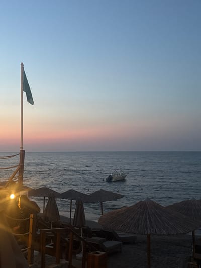 The image depicts a serene beach scene at sunset. The sky transitions from a clear blue at the top to soft hues of pink and orange near the horizon, indicating the time of day. The sea is calm, with gentle waves reflecting the twilight sky. In the water, a small boat is anchored, adding a touch of human activity to the natural landscape.

On the shore, several thatched umbrellas and beach chairs are set up, suggesting a resort or a public beach. The umbrellas are closed, perhaps due to the end of the day or to protect them from the evening air. The lighting suggests it's either early morning or late evening, with a warm glow from a light fixture visible in the foreground. The overall atmosphere is peaceful and relaxing, evoking a sense of tranquility often associated with sunsets. The location is identified as Chersonisos Municipal Unit, Greece, which aligns with the typical imagery of a Mediterranean beach. No text is visible in the image.