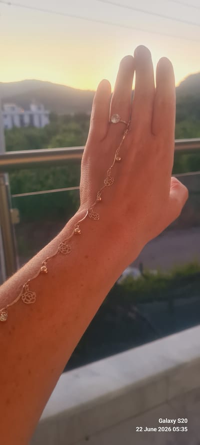 A person's left arm and hand are positioned in the foreground, with a delicate, gold-colored chain bracelet adorning their arm and hand. The bracelet features small, rounded charms and cut-out floral designs, connecting to a ring on the person's ring finger. The ring itself is simple, with a light-colored stone. The arm and hand are illuminated by warm, golden-hour sunlight, suggesting an outdoor setting. In the background, blurred green foliage and distant white buildings with red-tiled roofs are visible, set against a soft, hazy sky with a warm glow. This landscape is consistent with the scenery found in and around Dalaman, Turkey, particularly in residential or resort areas overlooking the natural terrain. The timestamp in the lower right corner indicates "Galaxy S20 22 June 2026 05:35," which is likely metadata from the device used to capture the image.