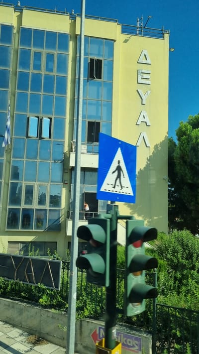 The image shows a building with a light yellow facade and numerous blue-tinted windows. The building has a balcony and a rooftop railing. Greek letters spelling "ΔΕΥΑΛ" are affixed to the facade of the building. A pedestrian crossing sign is visible in the foreground, mounted on a pole. Traffic lights are also present, with the top light illuminated red. A Greek flag is partially visible on the left side of the building. Dense green foliage and trees surround the building. A paved sidewalk is visible at the bottom of the image. The scene suggests an urban environment in Larissa, Greece, possibly in front of a public utility building.