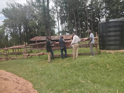 The image depicts a scene in a grassy, rural setting, likely in Idakho South ward, Kenya, based on provided information. Four African men are gathered near a rustic, wooden fence. To the right of the group, stands a large, black water tank. Behind the fence, there is a small, dilapidated structure with a corrugated metal roof. The background features a dense thicket of trees. The men are dressed in casual attire, and appear to be engaged in a discussion, possibly reviewing documents held by one of the men. The ground is covered in grass, with evidence of leaves or debris scattered across the surface. To the left of the grassy area is a dirt patch, possibly an exposed hillside. The weather appears overcast or cloudy, based on the diffused light.