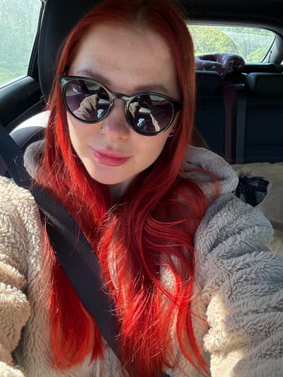 A selfie taken by a woman with red hair and sunglasses, likely inside a car. The woman is wearing a seatbelt and a fuzzy gray jacket. The location is not identifiable as Pokrovskoye, Russia from the given visual information.