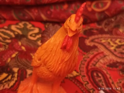 A close-up, low-angle shot shows an orange plastic toy chicken with red comb and wattle. The chicken is positioned centrally and is in focus, with the background appearing blurred. The background consists of a red fabric with a paisley-like pattern in shades of red, orange, and brown. A timestamp in the bottom right corner of the image reads "18.06.2026 11:42". The location is not specified in the image.