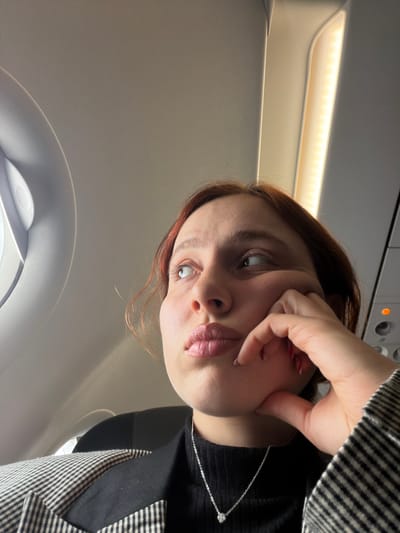 The image displays a female individual inside an airplane cabin. The scene is provided with the contextual location of Tuzla, Turkey. The subject, with reddish-brown hair, is positioned in the foreground, facing slightly right but looking upwards and to the left. Her left hand is resting with fingers lightly curled against her chin/jawline. She wears a black turtleneck or similar top, a black and white checkered blazer, and a silver-colored chain necklace with a small, light-colored pendant. Her fingernails are painted light red.

The immediate environment is an aircraft interior, specifically a window seat. A circular airplane window is visible on the upper left, framed by the light-colored interior paneling. The exterior view from the window is not discernible. The light-colored ceiling and overhead paneling are prominent. An illuminated strip of LED-like lights runs along the overhead panel on the right side of the image, casting a warm glow. An orange indicator light is visible on an overhead control panel on the far right. A dark headrest of an airplane seat is partially visible behind the person's right shoulder. No other individuals are visible, and there are no explicit interactions with other people.