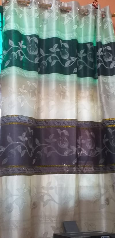 The image shows a close-up of a multi-colored curtain with a floral pattern. The curtain has horizontal stripes of green, cream, and dark purple. The floral pattern appears to be embossed or woven into the fabric and depicts branches with leaves and fruit-like shapes. The curtain is hung with grommets or rings, visible at the top. The lighting suggests an indoor setting, with a slight green hue cast on the upper part of the curtain, possibly from an external light source. There is a dark object, possibly a printer, visible in the lower right corner of the frame, partially obscured by the curtain. The overall impression is of a domestic or commercial interior. The location is identified as Sihanoukville, Cambodia.