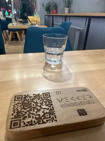 The image captures an indoor scene within the Vekker Kávéközösség cafe, located in Odorheiu Secuiesc, Romania. The vantage point is from a light-colored wooden table. A clear glass, approximately half-filled with water, stands on the table slightly right of center, casting a defined shadow with concentric light patterns. To the lower left, a rectangular wooden block, serving as a menu stand, features a laser-etched QR code on its left side. The right side displays the text "menu Box" and "VEKKER KÁVÉKÖZÖSSÉG," with a stylized 'N' (NFC symbol) below and a small '7' in a square above "VEKKER." In the midground, beyond the immediate table, dark teal upholstered chairs are visible around another light-colored wooden table. This secondary table holds a silver water bottle, a partially visible laptop with a yellow cover, and a stemmed glass containing a pink straw. The background shows light gray walls, a dark metal shelving unit topped with several potted plants, and a white radiator with a grid pattern. No individuals are present. The scene is illuminated by interior artificial lighting.
