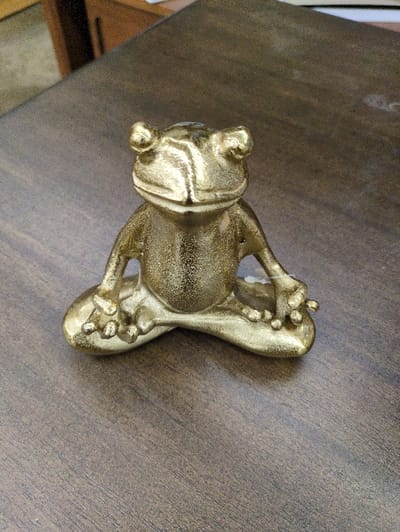 A gold-colored frog statue rests on a dark brown, textured surface. The frog is depicted in a relaxed pose, with its limbs spread out. The statue's surface is not smooth, displaying a grainy texture, possibly from the casting or finishing process. The lighting highlights the contours of the frog, emphasizing its form against the darker background.