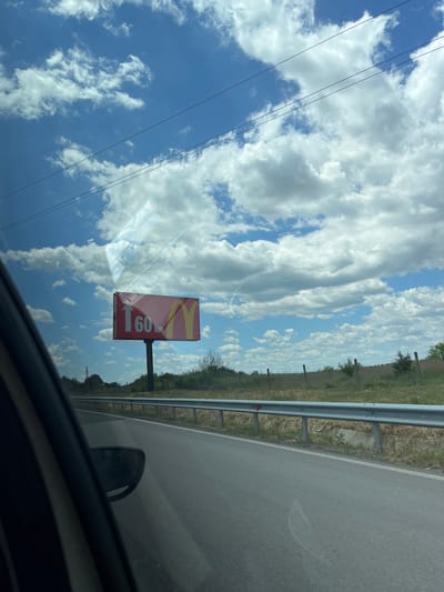 The image is a photograph taken from inside a vehicle looking out at a rural roadside scene. The sky is predominantly blue with scattered white, fluffy clouds. A red billboard is visible on the right side of the road, displaying the McDonald's logo and the text "60 km" with an upward-pointing arrow. A metal guardrail runs along the side of the asphalt road, separating it from a dry, grassy embankment. In the distance, trees and utility poles are visible under the expansive sky. The location is identified as Rupkite, Bulgaria, implying this scene is from a highway or road in that region. The overall impression is a bright, sunny day in a rural area.