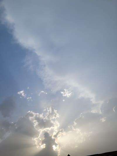 This image captures a sky scene in Nassarawan Eggon, Nigeria. The upper portion displays a clear to lightly overcast blue-gray sky, transitioning to a dense formation of clouds across the middle and lower sections. Bright crepuscular rays emanate from an intensely luminous, obscured light source, presumably the sun, located behind a heavy cloud bank towards the bottom-left quadrant. These rays extend upwards and outwards, illuminating cloud edges and creating stark contrasts between bright light and shadowed cloud masses. A dark, angular structure, consistent with a rooftop or architectural element, is visible in silhouette along the bottom edge of the frame. No individuals, animals, or specific identifiable objects are present beyond the described atmospheric and structural elements.