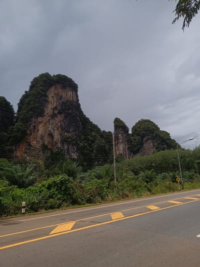 The image depicts a road near Khao Khram, Thailand. The road has yellow painted lane markings and speed bumps. Adjacent to the road is dense tropical vegetation with large, exposed rock formations rising out of the vegetation. The sky is overcast, suggesting an impending storm. Power poles and a streetlight are visible along the roadside. No people are present in the scene. A road warning sign with an arrow is visible on the shoulder of the road.